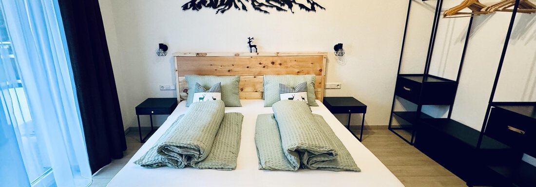 Ein modernes Schlafzimmer mit einem großen Holzbett und gemütlichen Kissen. An der Wand ist eine stilisierte Berglandschaft zu sehen, und es gibt Platz für Kleidung. - Appartement Bergglück - Umhausen