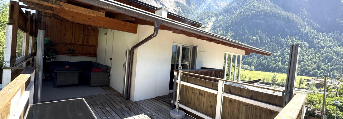 Eine moderne Terrasse mit Holzbelag und gemütlichen Sitzmöglichkeiten. Im Hintergrund sind hohe Berge und ein klarer blauer Himmel zu sehen. - Appartement Bergglück - Umhausen