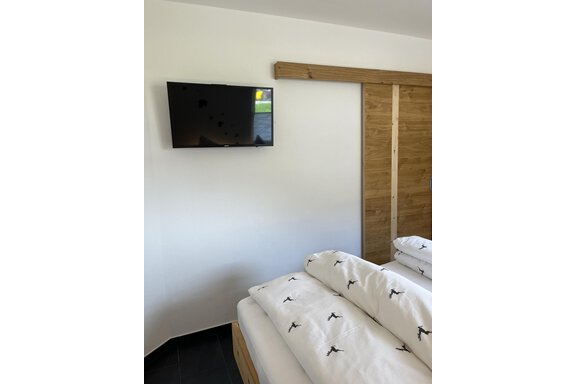 Ein modernes Schlafzimmer mit einem Fernseher an der Wand und einem komfortablen Bett. Der Raum hat eine minimalistische Gestaltung mit Holz- und weißen Farbtönen. - BLANKA'S Apart Auszeit - Umhausen