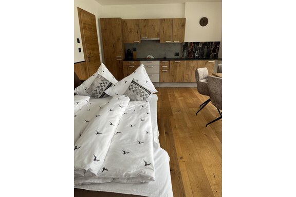 Ein modernes Zimmer mit einem Bett, das mit weißen Bettwäsche und Kissen dekoriert ist. Im Hintergrund ist eine kleine Küche mit Holzschränken sichtbar. - BLANKA'S Apart Auszeit - Umhausen