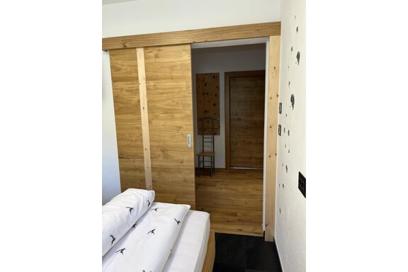 Ein Schlafzimmer mit einem Bett und Holzverkleidung. Im Hintergrund ist eine offene Tür zu einem weiteren Raum sichtbar. - BLANKA'S Apart Auszeit - Umhausen