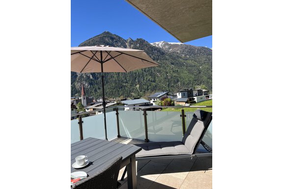 Eine moderne Terrasse mit einem Liegestuhl und einem Tisch. Im Hintergrund sind grüne Berge und ein blauer Himmel zu sehen. - BLANKA'S Apart Auszeit - Umhausen