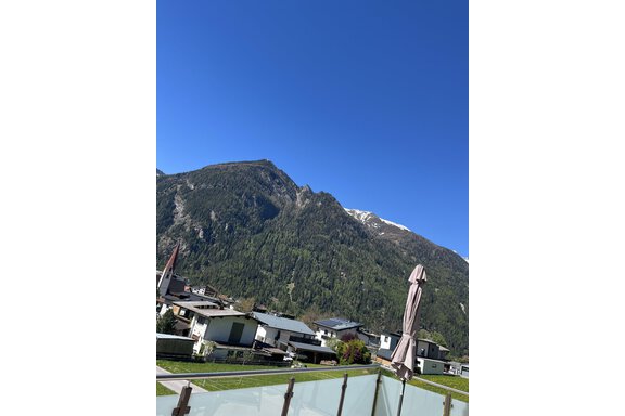 Eine malerische Berglandschaft mit einem klaren blauen Himmel. Im Vordergrund sind moderne Häuser und ein Sonnenschirm zu sehen. - BLANKA'S Apart Auszeit - Umhausen