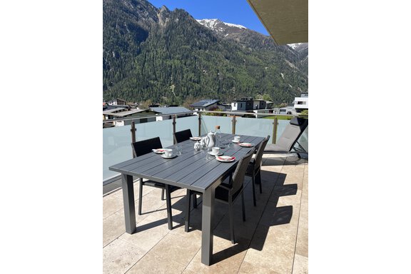 Ein moderner Essbereich auf einer Terrasse mit einem Tisch und Stühlen. Im Hintergrund sind grüne Berge und ein blauer Himmel zu sehen. - BLANKA'S Apart Auszeit - Umhausen