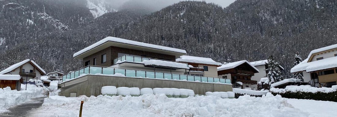 Ein modernes Haus in einer verschneiten Landschaft. Im Hintergrund sind hohe Berge und ein bewölkter Himmel zu sehen. - BLANKA'S Apart Auszeit - Umhausen