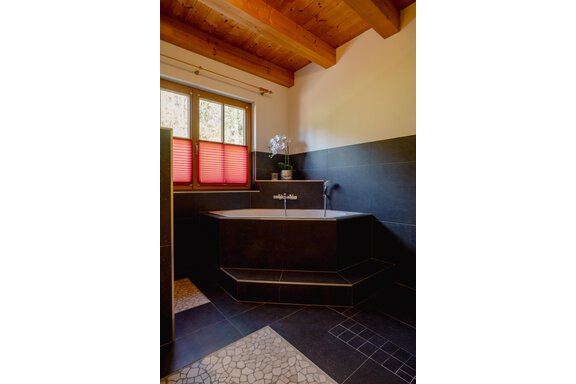 A modern bathroom with an elegant bathtub and dark wall tiles. Large windows with red blinds let in plenty of light. - Chalet Carla - Umhausen
