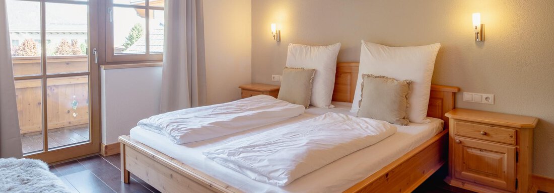A cozy bedroom with a wooden double bed and white bedding. Large windows let in plenty of daylight and offer a view outside. - Chalet Carla - Umhausen