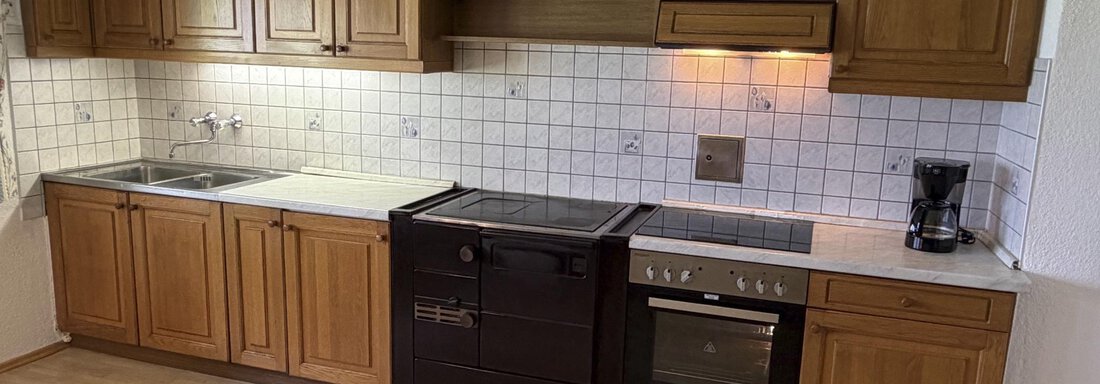 A modern kitchen with wooden cabinets and white tiles. It includes a cooktop, an oven, and a coffee machine. - Ferienhaus am Spielroan - Umhausen