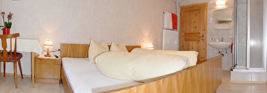 A cozy room with a double bed and bright pillows. In the background, there is a small bathroom and a table with a chair. - Ferienhaus am Spielroan - Umhausen