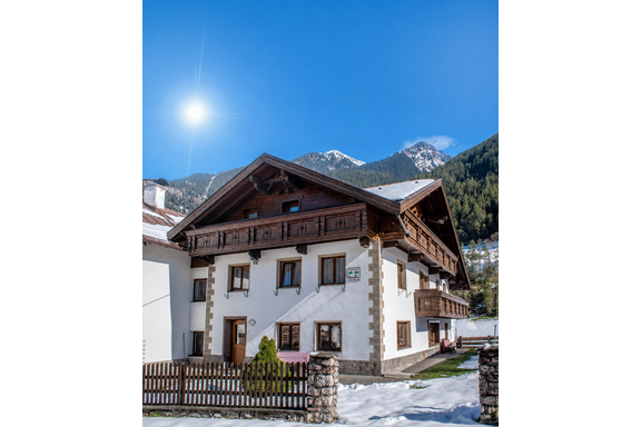 A traditional chalet in the Alps with a wood-decorated facade. In the background, snow-covered mountains stretch under a clear blue sky. - Ferienhaus am Spielroan - Umhausen