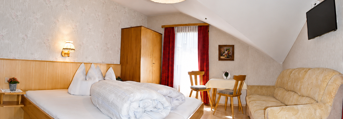 A cozy bedroom with a double bed, a couch, and a small dining area. Bright walls and large windows create a friendly atmosphere. - Ferienhaus am Spielroan - Umhausen