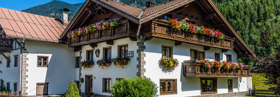 A picturesque house in alpine style with colorful flower boxes. In the background, green mountains and a radiant blue sky are visible. - Ferienhaus am Spielroan - Umhausen