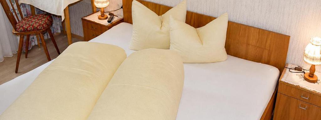 A simple bedroom with a double bed and two large yellow pillows. Next to the bed are nightstands with lamps and a chair at the table. - Ferienhaus am Spielroan - Umhausen