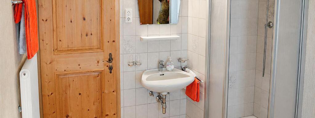 A bright bathroom with a shower niche and a small sink. The walls are lined with white tiles, and there is a wooden door. - Ferienhaus am Spielroan - Umhausen