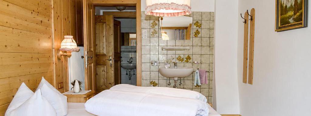 A cozy bedroom with a large bed and wooden walls. In the background, there is a bathroom with tiles and a sink. - Ferienhaus am Spielroan - Umhausen