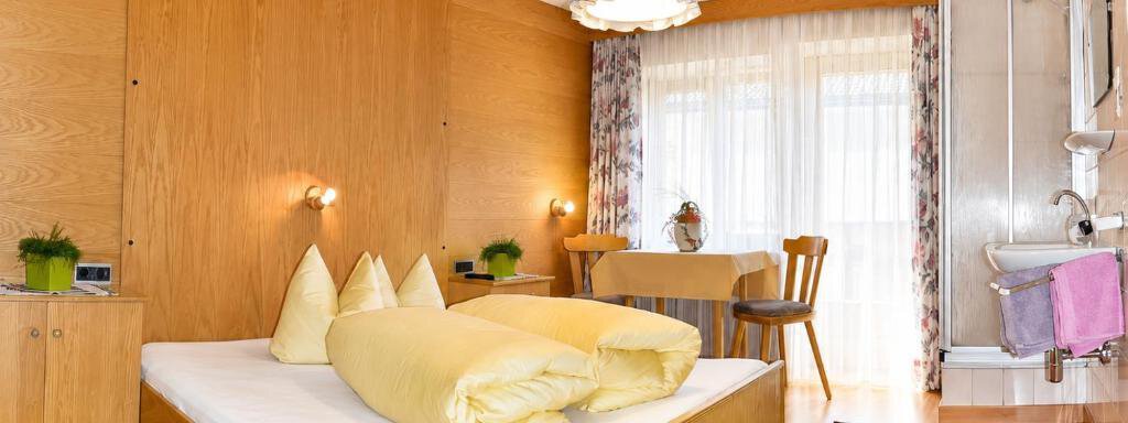 A cozy room with wooden walls and a large window. It is furnished with a double bed, a table, and chairs. - Ferienhaus am Spielroan - Umhausen