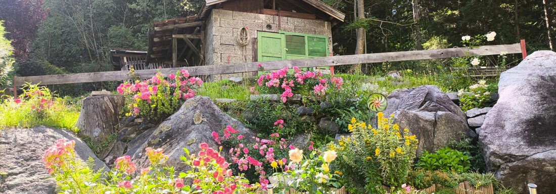 A cozy wooden house surrounded by colorful flowers and large stones. The garden is green and surrounded by trees, creating a calm atmosphere. - Ferienwohnung Martha - Umhausen