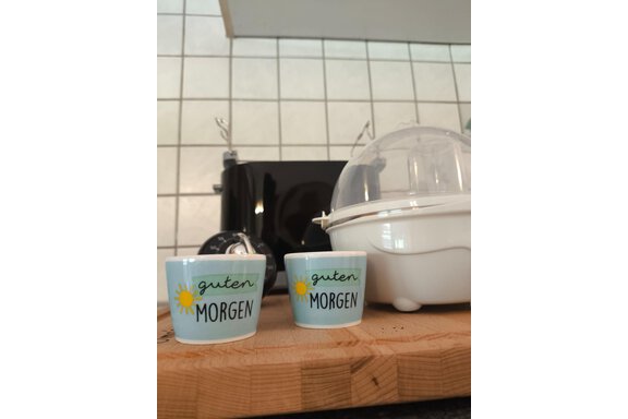 Two cups with the inscription "good MORNING" are placed on a wooden board. In the background, a black toaster and a juicer can be seen. - Ferienwohnung Martha - Umhausen