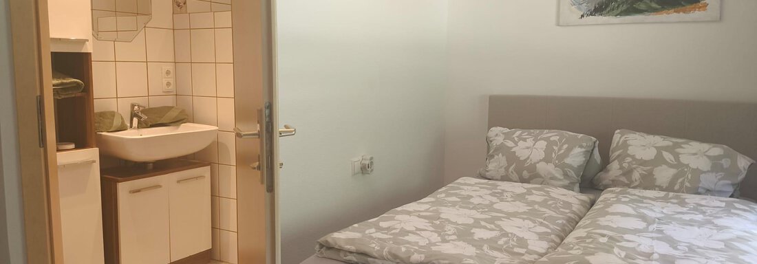 A cozy bedroom with a double bed and fresh bed linens. In the background, a bathroom with a modern sink is visible. - Ferienwohnung Martha - Umhausen
