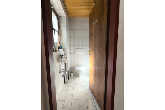 A small bathroom with white tiles and a toilet. The door is made of wood and there is a washbasin with a mirror. - Ferienwohnung Martha - Umhausen