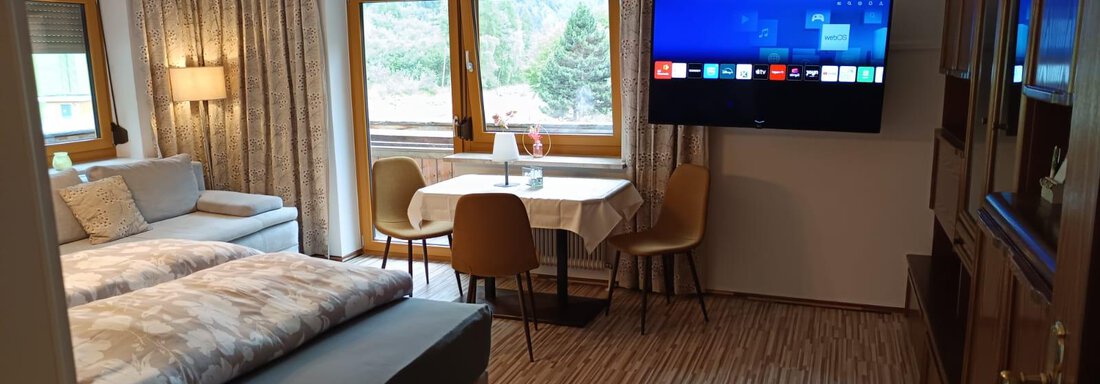 A cozy room with a double bed, a seating area, and a table. A large television is mounted on the wall, and there are many windows overlooking the greenery. - Ferienwohnung Martha - Umhausen