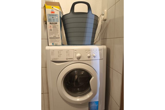 A washing machine is in a small room. On top of the machine is a gray laundry basket. - Ferienwohnung Martha - Umhausen
