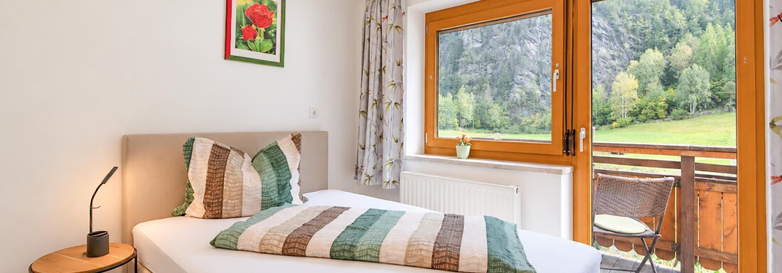 A cozy bedroom with a large window that overlooks a green landscape. The bed is simple and decorated with colorful pillows. - Ferienwohnung Martha - Umhausen