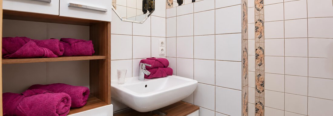 A modern bathroom with white tiles and a sink. Pink towels are neatly stacked and a shower area is visible. - Ferienwohnung Martha - Umhausen