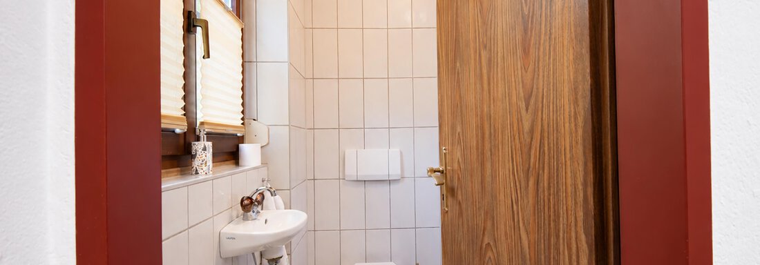 A small bathroom with tiled walls and a wooden ceiling. There is a sink, a toilet, and a window for daylight. - Ferienwohnung Martha - Umhausen