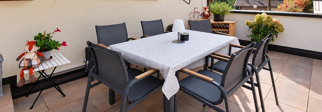 An inviting terrace area with a large table and chairs. In the background, there are plants and a picturesque mountain landscape. - Ferienwohnung Martha - Umhausen