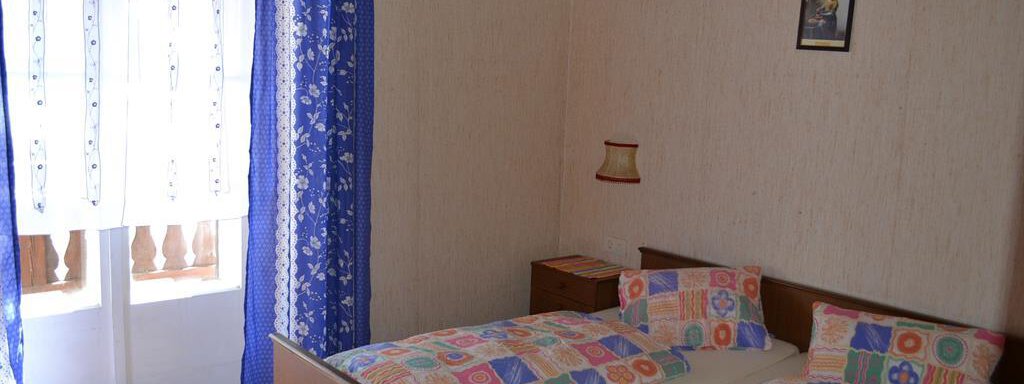 A simple bedroom with two beds and colorful bedspreads. The windows are decorated with blue curtains, and there is a small bedside table. - Appt. Garni Anny - Vent
