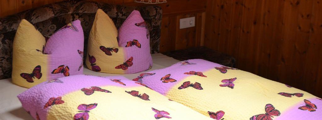 A cozy bed with a blanket and pillows in yellow and purple, adorned with butterfly patterns. The wooden walls create a warm, inviting atmosphere. - Appt. Garni Anny - Vent