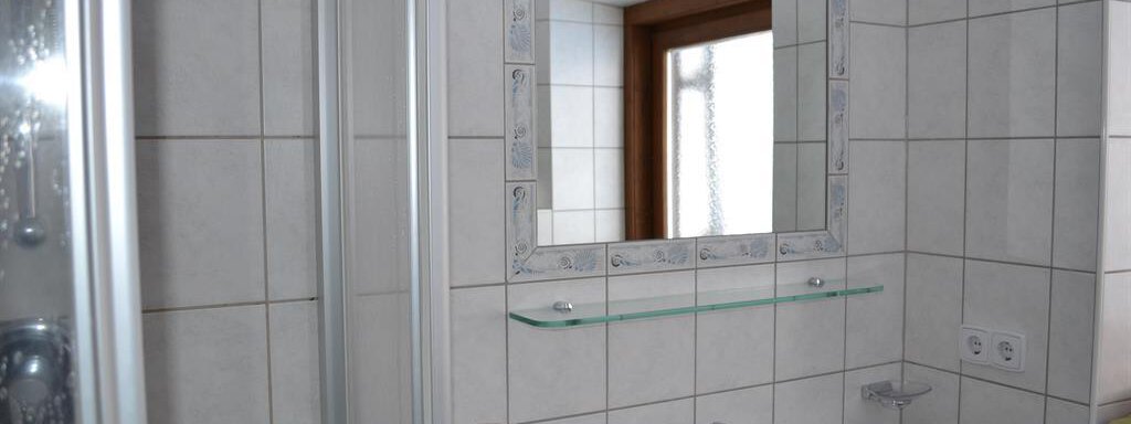 A bright bathroom with white tiles and a large mirror. There is a sink and a shower cabin. - Appt. Garni Anny - Vent