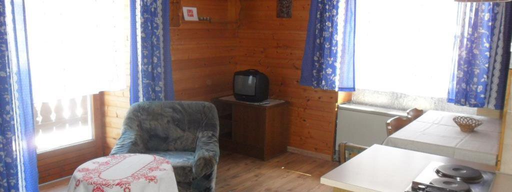 A cozy living room with wooden walls and large windows. There is an armchair, a small table, and a kitchenette in the background. - Appt. Garni Anny - Vent