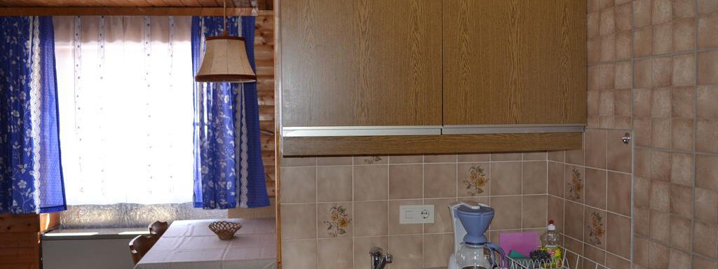 A small, cozy kitchen with wooden furniture and a sink. The window is decorated with blue curtains, and there is a cooking area as well as a coffee machine. - Appt. Garni Anny - Vent