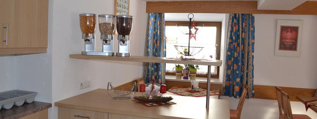 A bright kitchen with modern wood design and a spacious dining area. On the table are various snacks in containers, and colorful curtains enhance the cozy atmosphere. - Appt. Garni Anny - Vent
