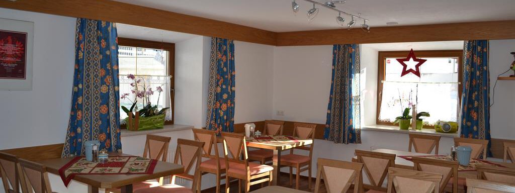 A cozy room with wooden tables and chairs. The windows are decorated with colorful curtains, and there are orchids on the windowsills. - Appt. Garni Anny - Vent