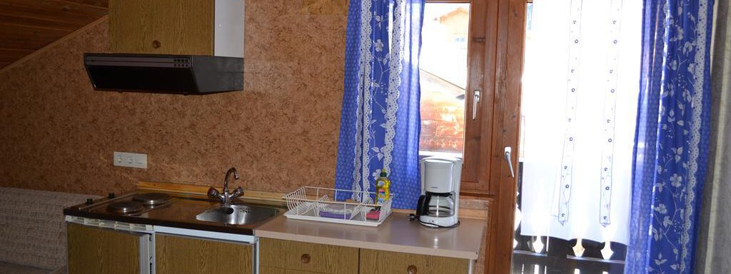 A small, cozy kitchenette with a stove, a sink, and a coffee maker. Blue curtains are hanging next to a window, allowing plenty of daylight in. - Appt. Garni Anny - Vent