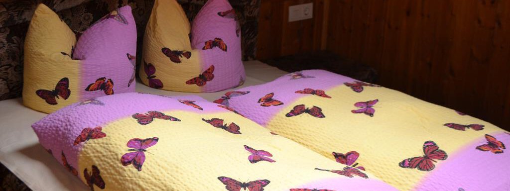 A cozy bed with colorful blankets and pillows displaying butterflies. The bedroom environment has a warm, rustic atmosphere. - Appt. Garni Anny - Vent