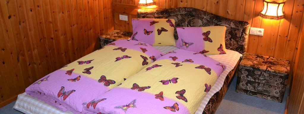 A cozy bedroom with a double bed and colorful bedspreads featuring a butterfly pattern. The walls are made of wood, and two lamps provide pleasant lighting. - Appt. Garni Anny - Vent