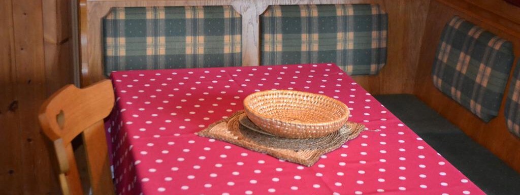 A rustic dining table with a pink tablecloth with white dots. The table is decorated with a woven basket and a natural mat, surrounded by a cozy seating area. - Appt. Garni Anny - Vent