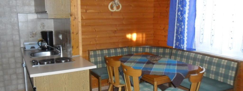 A cozy kitchen area with wooden walls and a dining table. The bench is covered with checked fabric, and the light falls through a window with blue curtains. - Appt. Garni Anny - Vent