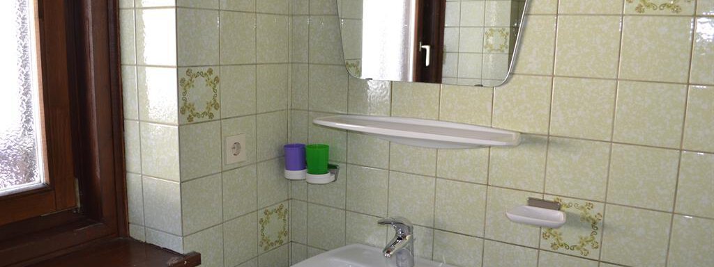 A small bathroom with green tiles and a sink. There is a window and a mirror above the sink. - Appt. Garni Anny - Vent