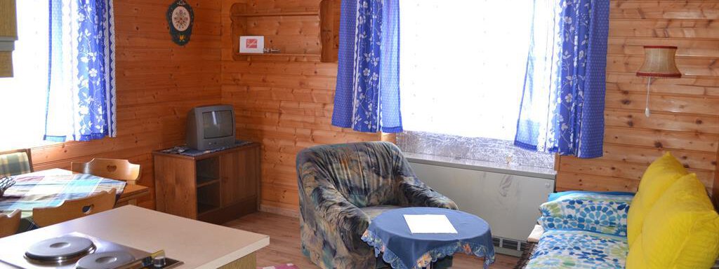 A cozy living space with wooden walls and large windows. There is a sofa, a table, and a small kitchen corner. - Appt. Garni Anny - Vent