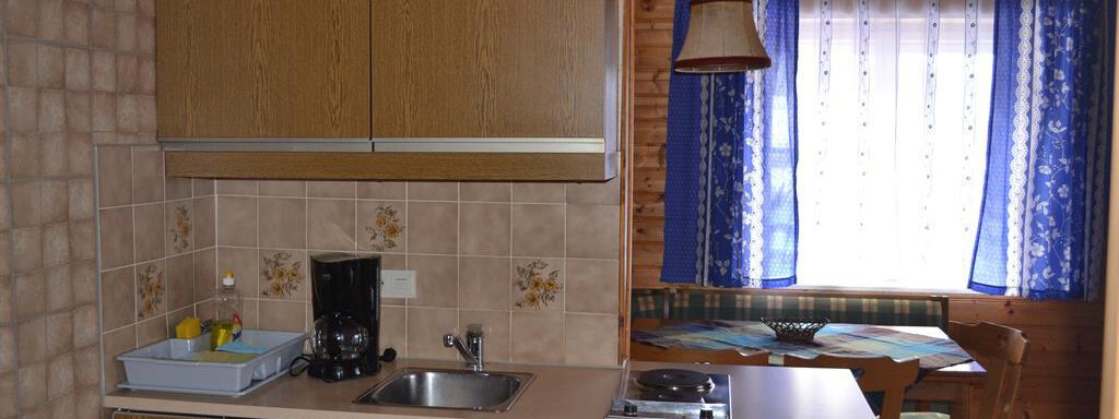 A small kitchen with wooden cabinets and a sink. In the background, there is a table and chairs, as well as a window with blue curtains. - Appt. Garni Anny - Vent