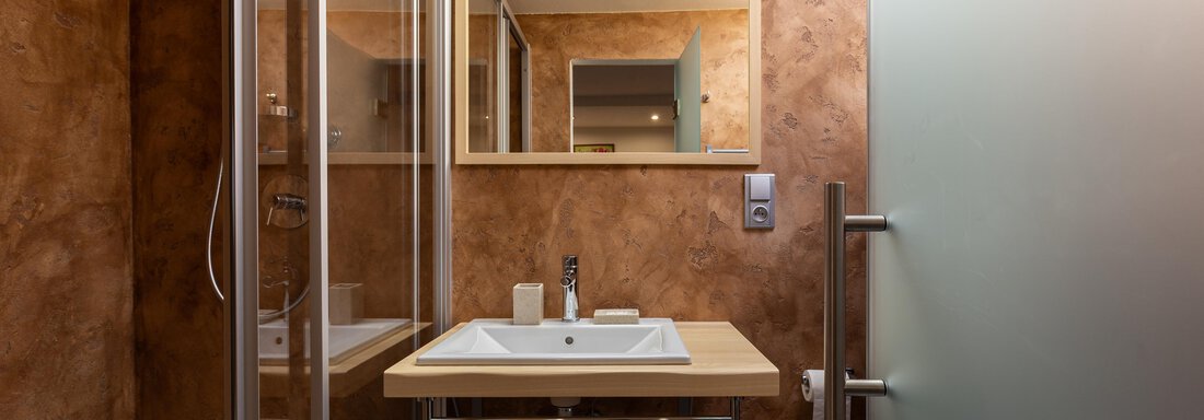 A modern bathroom with a clear shower, a sink, and a large mirror. The walls are in warm brown tones, and there is a minimalist decor. - Garni Stefani - Vent