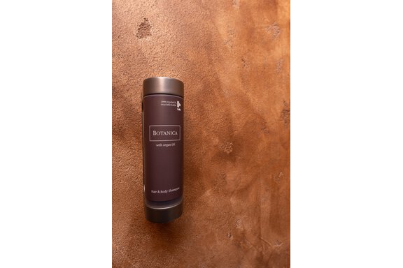 A bottle of shampoo and shower gel is placed on a rustic surface. The product is labeled "Botanica" and contains argan oil. - Garni Stefani - Vent