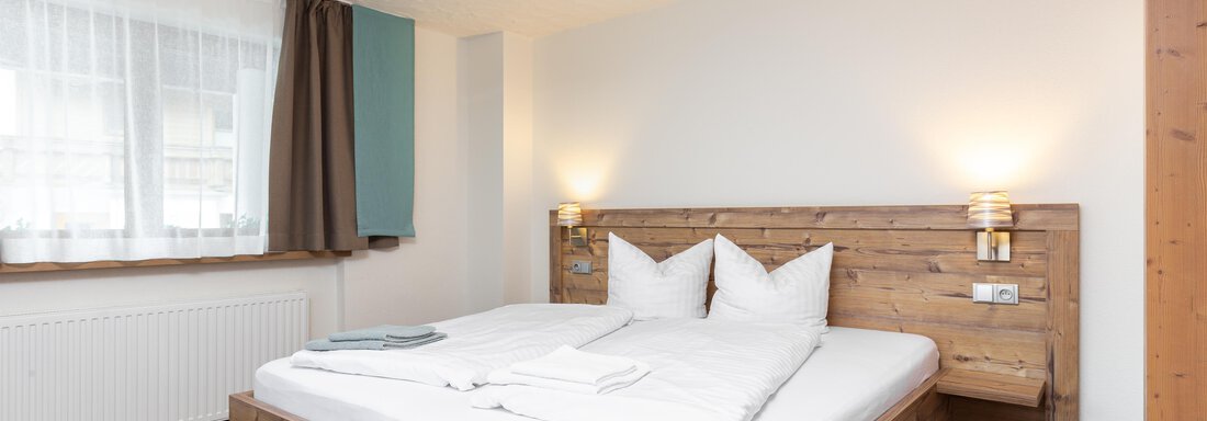 A cozy bedroom with a wooden double bed and two pillows. The room is bright with large windows and a modern, simple design. - Garni Stefani - Vent