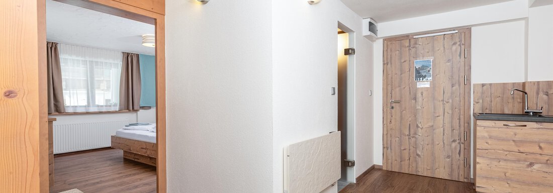 A bright, modern room with a wooden floor and warm colors. In the background, a bedroom and a small kitchen corner are visible. - Garni Stefani - Vent
