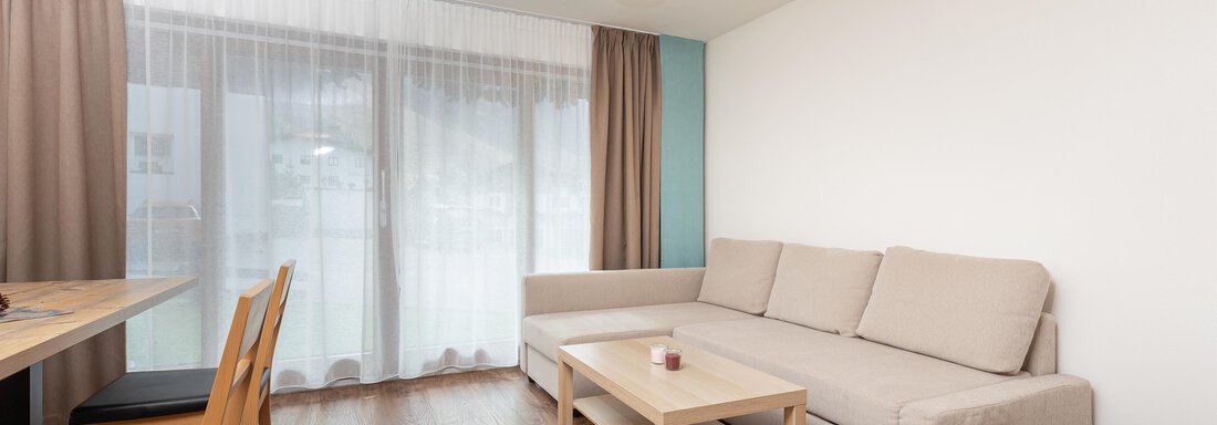 A modern living room with a bright sofa and a table for four people. Large windows provide plenty of daylight and a view outside. - Garni Stefani - Vent
